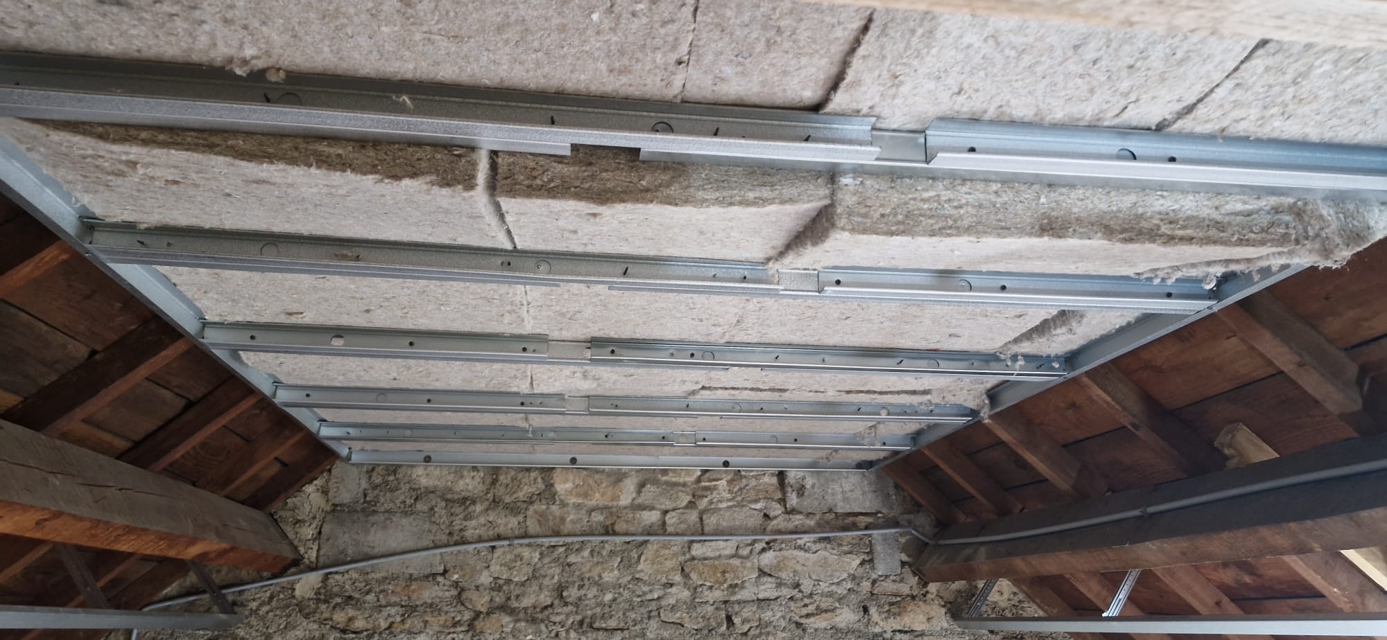 Ossature métallique de plafond fixée sous charpente avec isolation en laine minérale entre murs en pierre